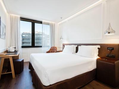 Helles Hotelzimmer mit großem Bett, Holzfußboden, Schreibtisch und Fenster mit Blick auf Gebäude.