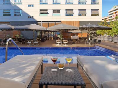 Schwimmbad mit Sonnenschirmen, Liegestühlen und einem Tisch mit Getränken vor einem Hotelgebäude.