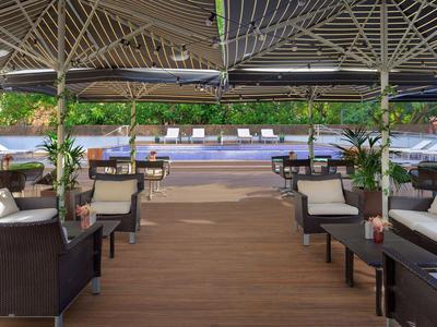 Überdachte Terrasse mit Holzboden, braunen Sesseln, weißen Sofas und Pflanzen am Straßenrand.