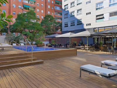 Moderne Terrasse mit Holzdeck, Liegen, Stufen und Bäumen vor mehrstöckigen Gebäuden.