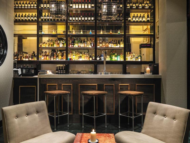 Gemütliche Bar mit zwei beigen Sesseln, kleinem Tisch, schwarzen Hockern und beleuchteten Regalen.