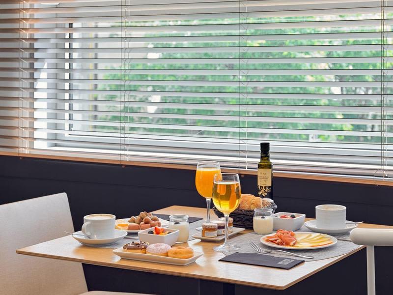 Kleiner Tisch mit Frühstücksgedeck, zwei Gläser Orangensaft, Weinflasche und Sitzplätze am Fenster mit Jalousien.
