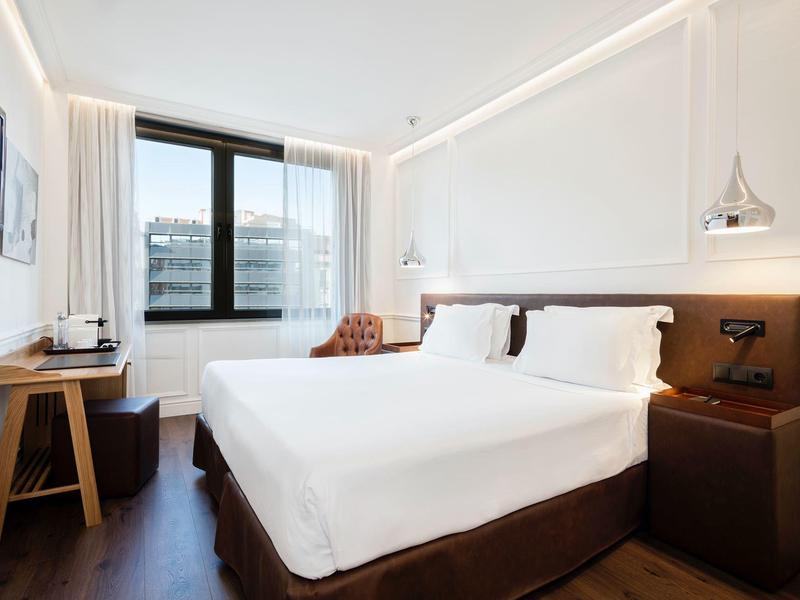 Helles Hotelzimmer mit großem Bett, Holzfußboden, Schreibtisch und Fenster mit Blick auf Gebäude.