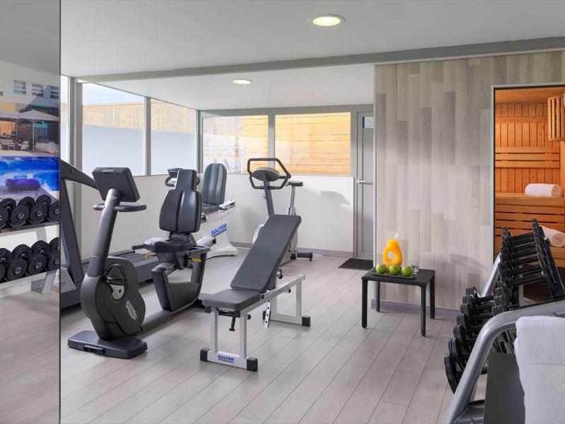 Moderner Fitnessraum mit Trainingsgeräten, Hanteln und Sauna, hell und geräumig gestaltet.