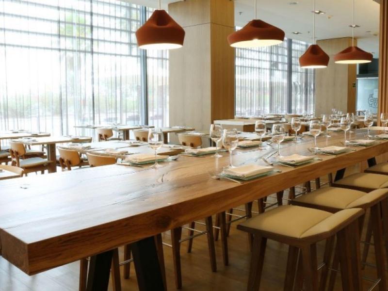 Großer Raum mit langem Holztisch, Hockern, großen Fenstern und moderner Beleuchtung.