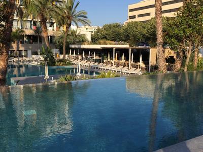 Gran piscina exterior con tumbonas y sombrillas junto a palmeras y edificios del hotel.