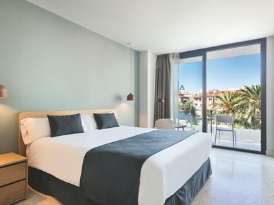 Helles Hotelzimmer mit Doppelbett, Nachttischen und Balkon mit Sicht auf Palmen.