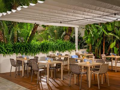 Moderner Außenbereich eines Hotels mit Holzboden und vielen Stühlen und Tischen unter einem weißen Sonnenschutz.