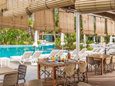 Offener Essbereich mit Holztischen und Stühlen neben einem Pool mit Liegestühlen und Pflanzen.