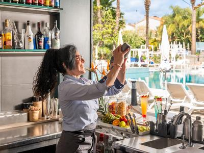 Eine Barkeeperin mischt Cocktails an einer belebten Hotelbar mit Pool im Hintergrund.