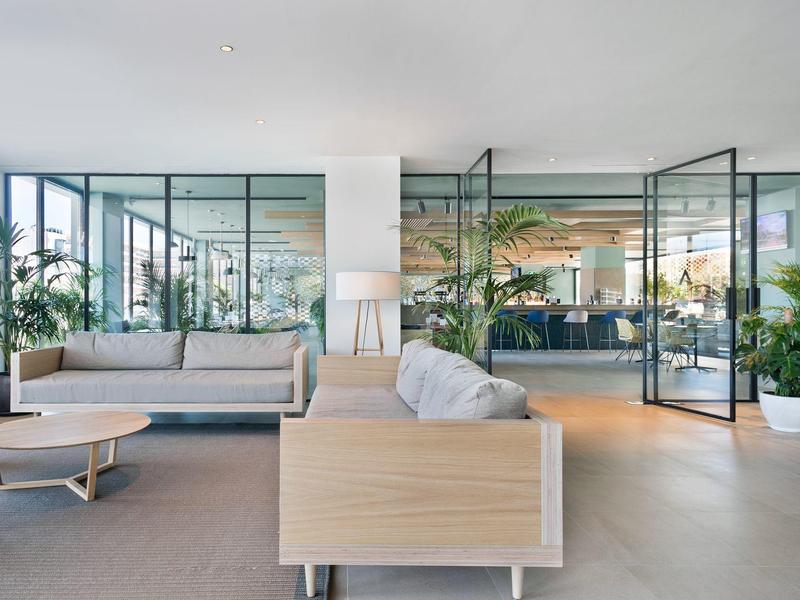 Área de salón de hotel luminosa y moderna con sofá, muebles de madera y grandes paredes de vidrio hacia un restaurante.