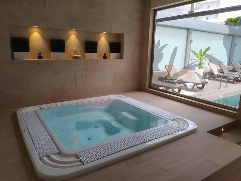 Binnenzwembad met bubbelbad en uitzicht op buitenzwembad met ligbedden en planten.