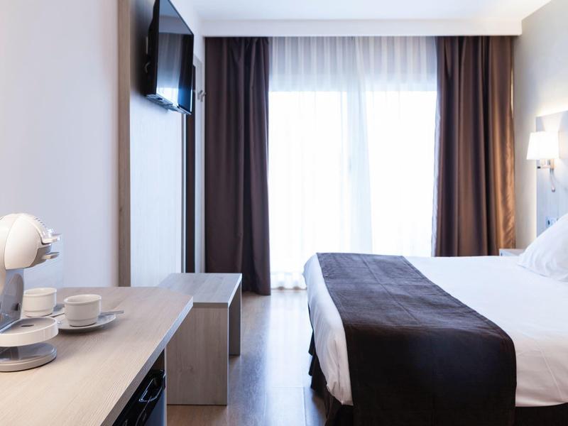 Lichte hotelkamer met groot bed, tv, koffiezetapparaat en gordijnen voor balkon.