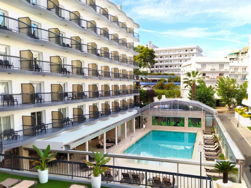 Hotel met buitenzwembad, ligstoelen, balkons en groene planten onder een blauwe lucht.