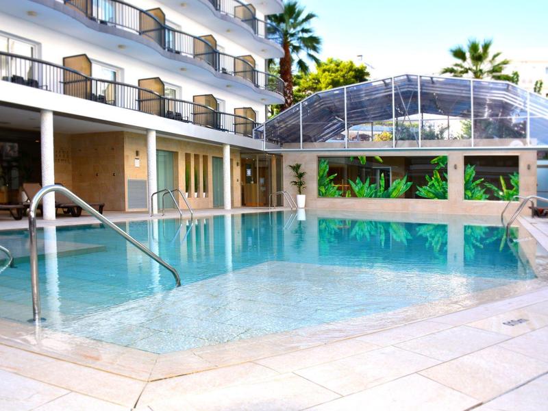 Buitenzwembad van hotel met helder water en trappen, omgeven door een meerlaags gebouw en palmbomen.