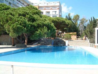 Helderblauw zwembad met waterval en grote bomen bij een hotelgebouw.