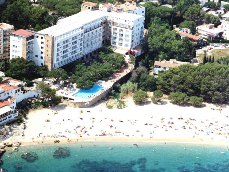 Hotel met zwembad aan een wit zandstrand en helder turquoise water, omgeven door bomen.