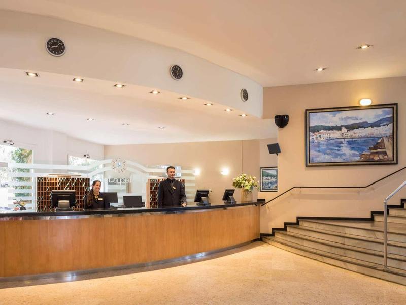 Lichte hotelreceptie met lange houten balie, receptiemedewerkers en een schilderij aan de muur.