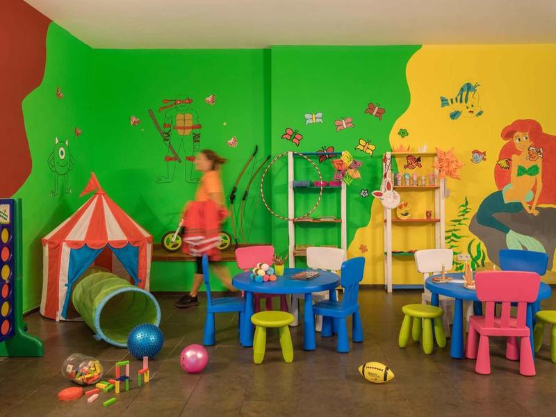 Kleurrijke speelruimte voor kinderen met tafels, stoelen, tunnel en muurschilderingen.