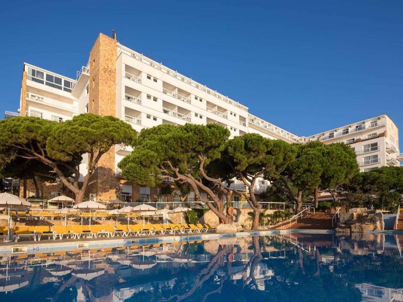 Groot hotel met buitenzwembad, parasols en schaduwgevende bomen bij klare lucht.