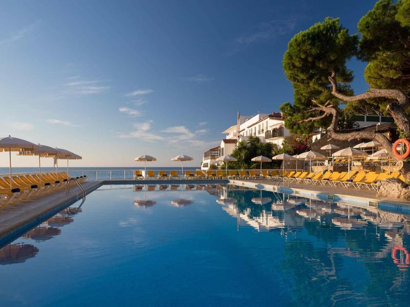 Groot zwembad met gele ligstoelen en parasols voor een hotel aan zee.