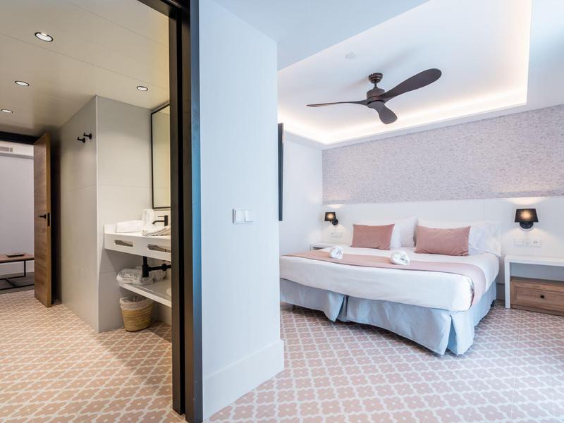 Moderní hotelový pokoj s manželskou postelí, stropním ventilátorem a světlou koupelnou.