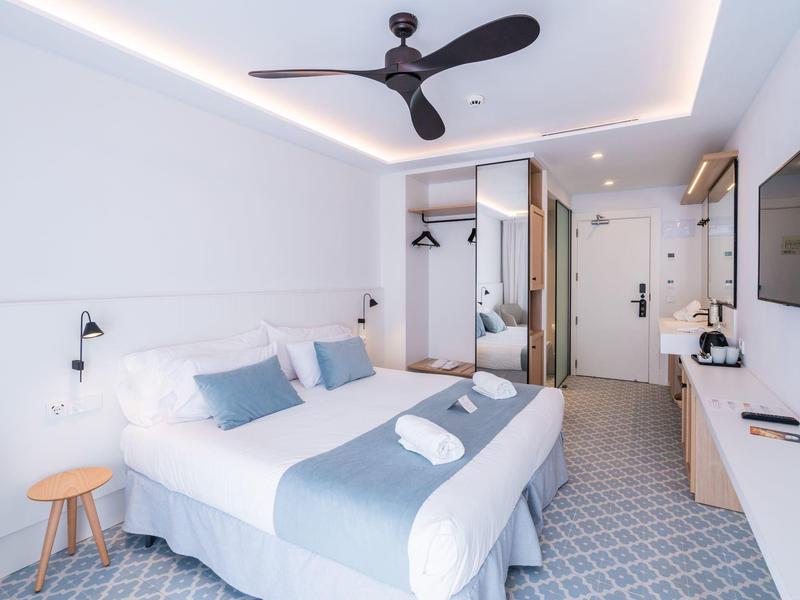 Moderní hotelový pokoj s manželskou postelí, stropním ventilátorem a jednoduchým zařízením.