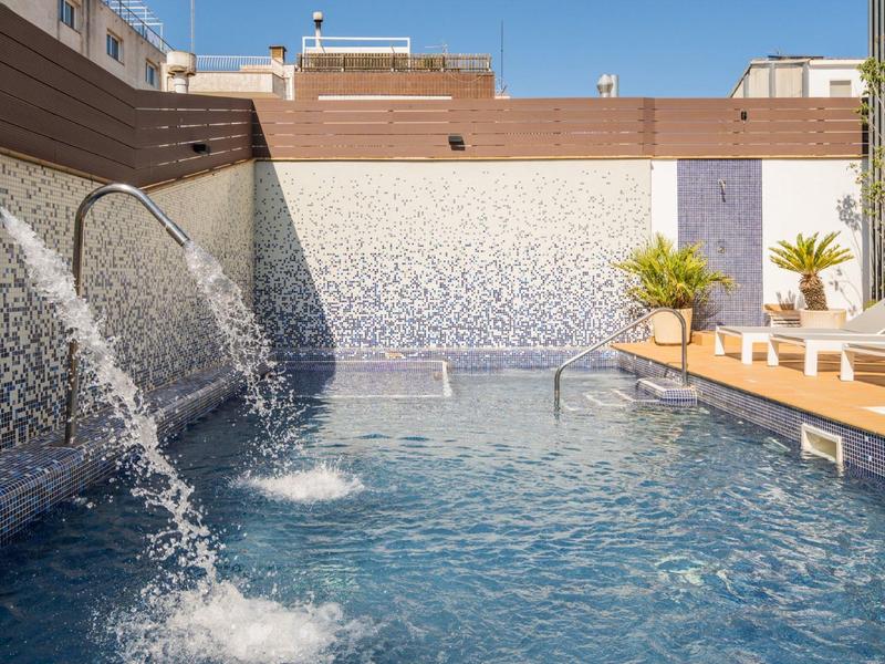 Venku bazén s vodními tryskami, lehátka a rostliny po stranách.