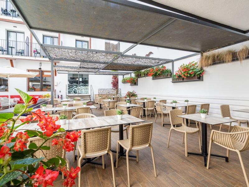 Moderní hotelová terasa s jídelními stolky, proutěnými židlemi a červenými květinami pod částečnou střechou.