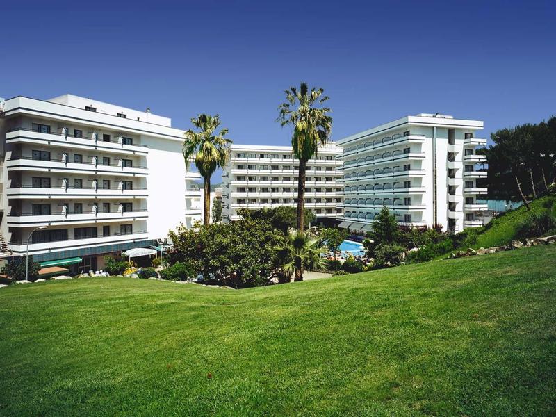 Due grandi hotel bianchi con balconi separati da palme e una piscina sotto un cielo azzurro limpido.