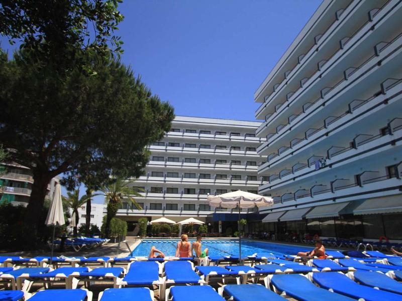 Area piscina all'aperto con lettini blu, ombrelloni e moderni edifici dell'hotel sotto un cielo sereno.