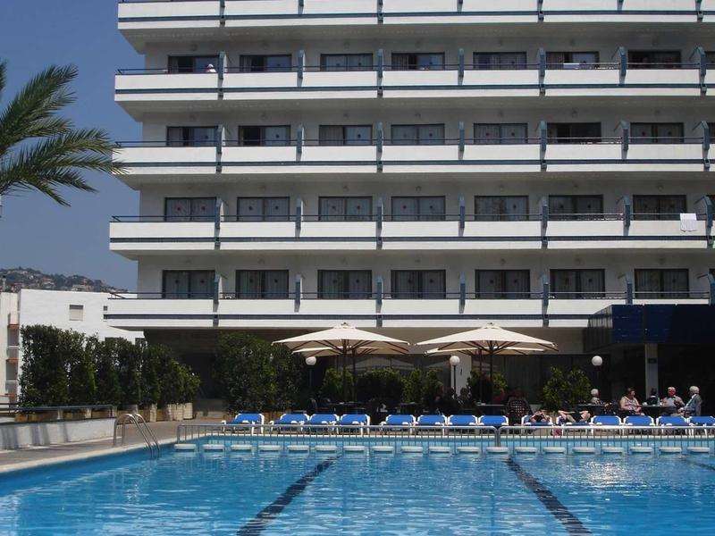 Hotel moderno con balconi che si affacciano su una grande piscina all'aperto sotto un cielo azzurro limpido.