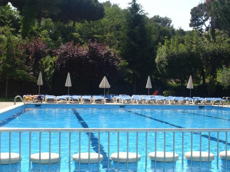 Piscina all'aperto con acqua blu, lettini, ombrelloni bianchi e alberi intorno.