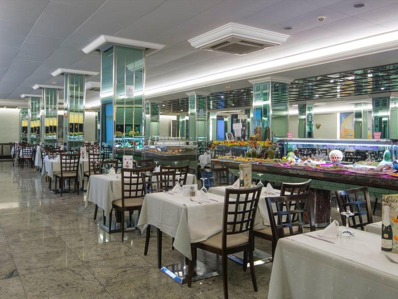 Großer Speisesaal mit weißen Tischdecken und vielen Stühlen, heller Beleuchtung und Spiegeln an der Wand.