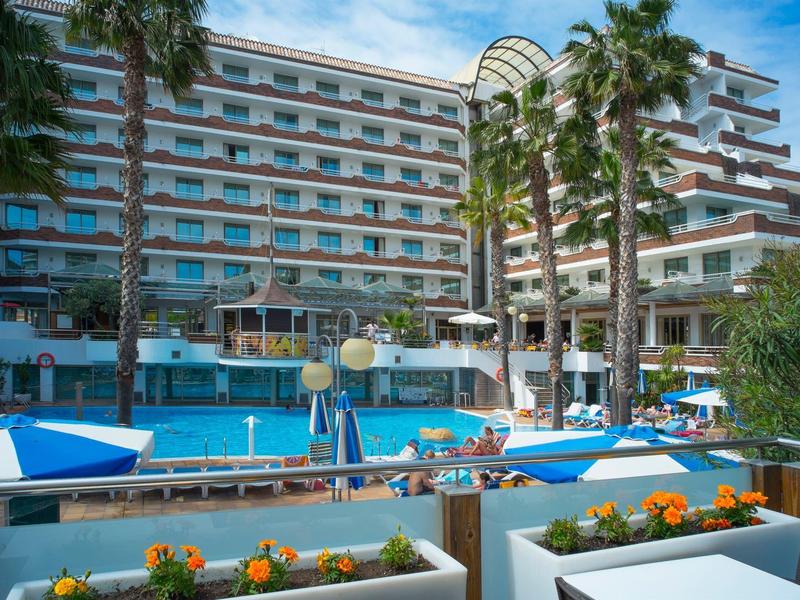 Moderner Hotelkomplex mit Swimmingpool, Sonnenschirmen und Palmen in einer sonnigen Umgebung.