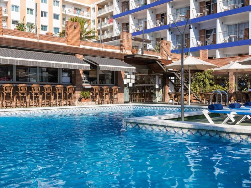 Outdoor hotel pool with sun loungers and umbrellas near a bar area under a building.