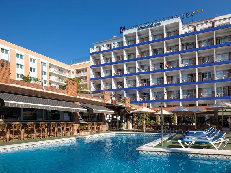 Outdoor pool with lounge chairs in front of a multi-story hotel under a clear blue sky.