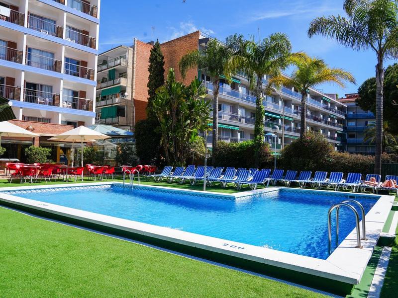 Outdoor swimming pool with sun loungers in front of multi-story hotel buildings and palm trees on a sunny day.