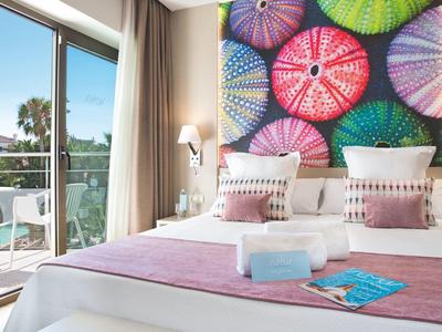 Světý hotelový pokoj se dvoulůžkem, barevným uměleckým dílem a balkonem s výhledem na palmy.