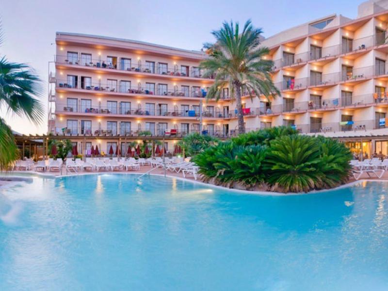 Velký hotelový bazén obklopený palmami s vícepodlažní hotelovou budovou v pozadí.