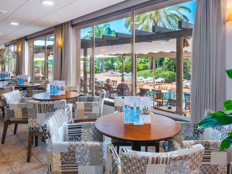 Prosvětlená restaurace s proutěnými židlemi a velkými okny s výhledem na terasu a palmy.