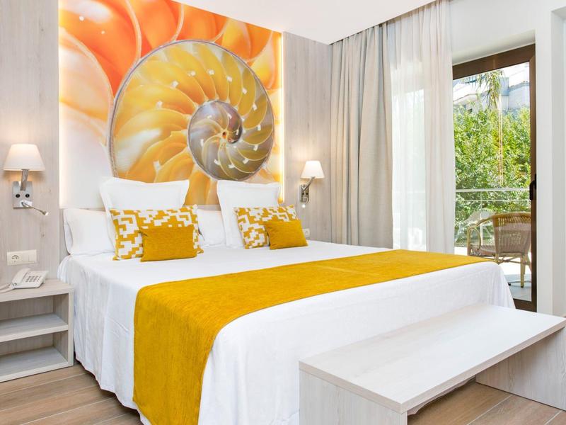 Světý hotelový pokoj s velkou postelí, žlutým přikrývkou a obrazem mušle na stěně.