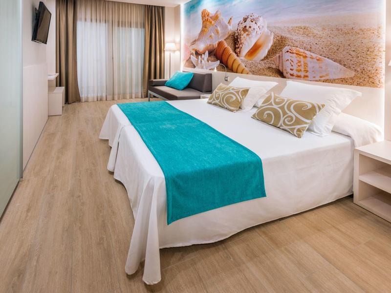 Světle hotelový pokoj s manželskou postelí, tyrkysovým přehozem a nástěnným obrazem moře.
