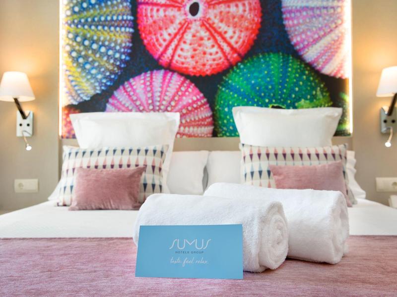 Moderní hotelový pokoj s bílou postelí, růžovými doplňky a barevným uměleckým dílem nad čelem postele