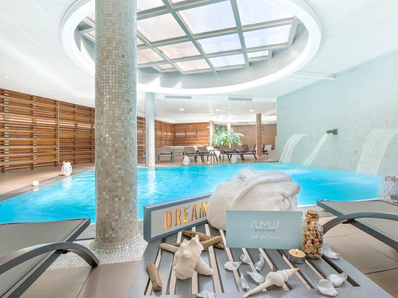 Moderní vnitřní bazén s masivním sloupem, lehátky a spa doplňky v světlém prostředí.