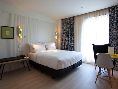 Modernes Schlafzimmer mit großem Bett, schwarzen Vorhängen, Schreibtisch und Stühlen bei Tageslicht.