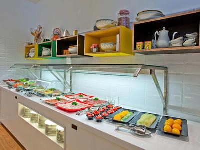 Buffet mit vielfältigen Frühstücksspeisen, bunte Regale mit Geschirr und helle Kacheln im Hintergrund.