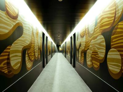 Langer Gang mit dunklen Wänden, verziert mit großen, gelben, organischen Mustern und hell beleuchtetem Boden.