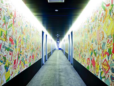 Langer Flur mit farbenfrohen, abstrakten Wandgemälden und grauem Bodenbelag, beleuchtet von Deckenlichtern.