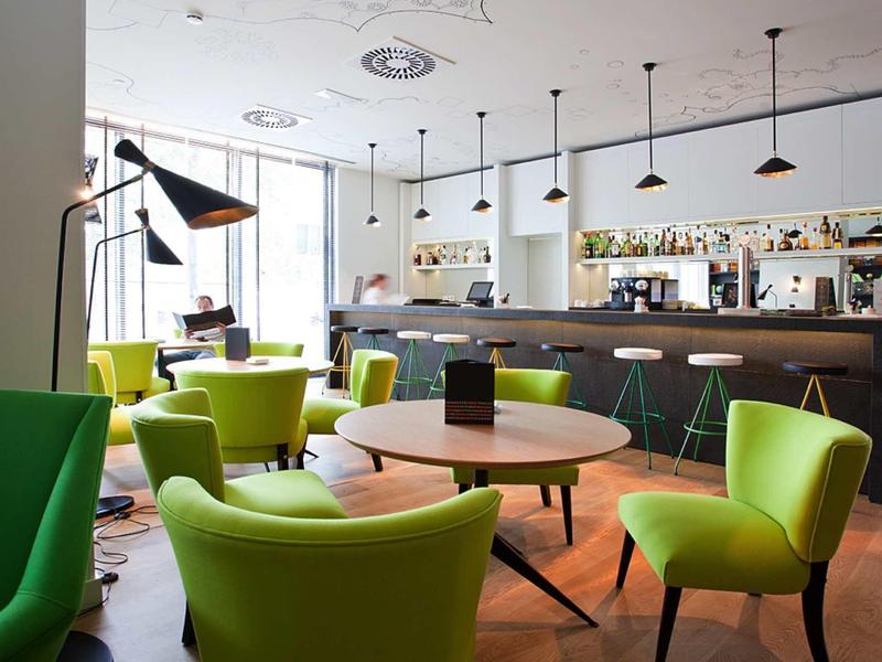 Modernes Café mit grünen Sesseln, runden Holztischen und weißer Bar unter Hängelampen.